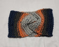 A handmade knitted twist-front ear warmer in a variegated pattern of navy blue, charcoal gray, rose pink, and light gray, laid flat on a white fabric background.