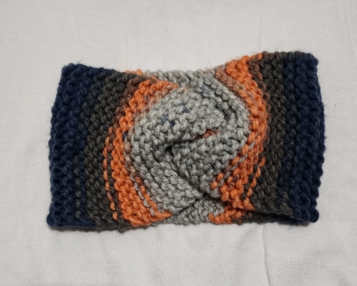 A handmade knitted twist-front ear warmer in a variegated pattern of navy blue, charcoal gray, rose pink, and light gray, laid flat on a white fabric background.