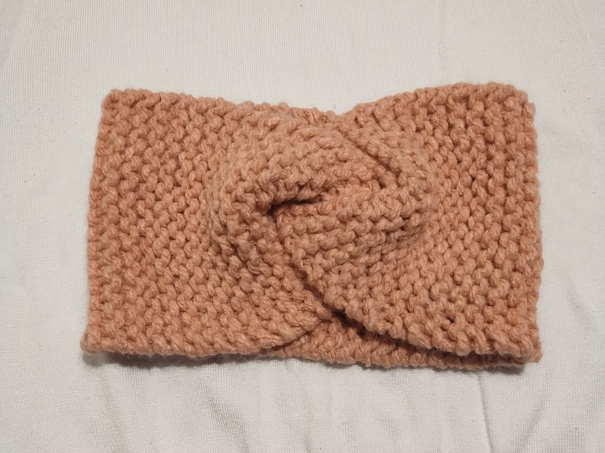 A chunky knit twisted headband in a dusty rose color, laid flat on a white fabric background.